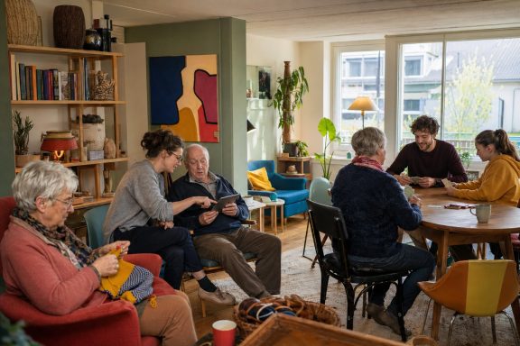 Zeit miteinander · Konzeptvisualisierung Unverbindliche KI-Konzeptvisualisierung eines wohnlichen Treffpunkts mit älteren und jüngeren Nachbarn bei Handarbeit, Kartenspiel und Tablet-Hilfe.
