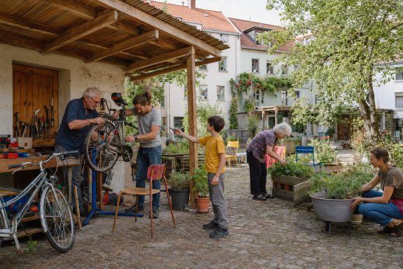 Werkstatt und Garten · Konzeptvisualisierung Unverbindliche KI-Konzeptvisualisierung einer offenen Hofwerkstatt mit Fahrradreparatur, Hochbeeten und generationenübergreifender Alltagshilfe.